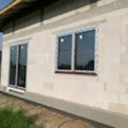 Nowo zamontowane okna z brązowymi ramami w surowym stanie budynku z białego betonu komórkowego. Widoczne szare rolety zewnętrzne częściowo opuszczone. Budowa w trakcie realizacji, widoczne elementy...