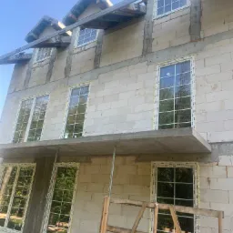 Wielopiętrowy budynek w trakcie budowy z zamontowanymi oknami z białymi ramami, widoczne szare bloki konstrukcyjne i drewniane rusztowanie.