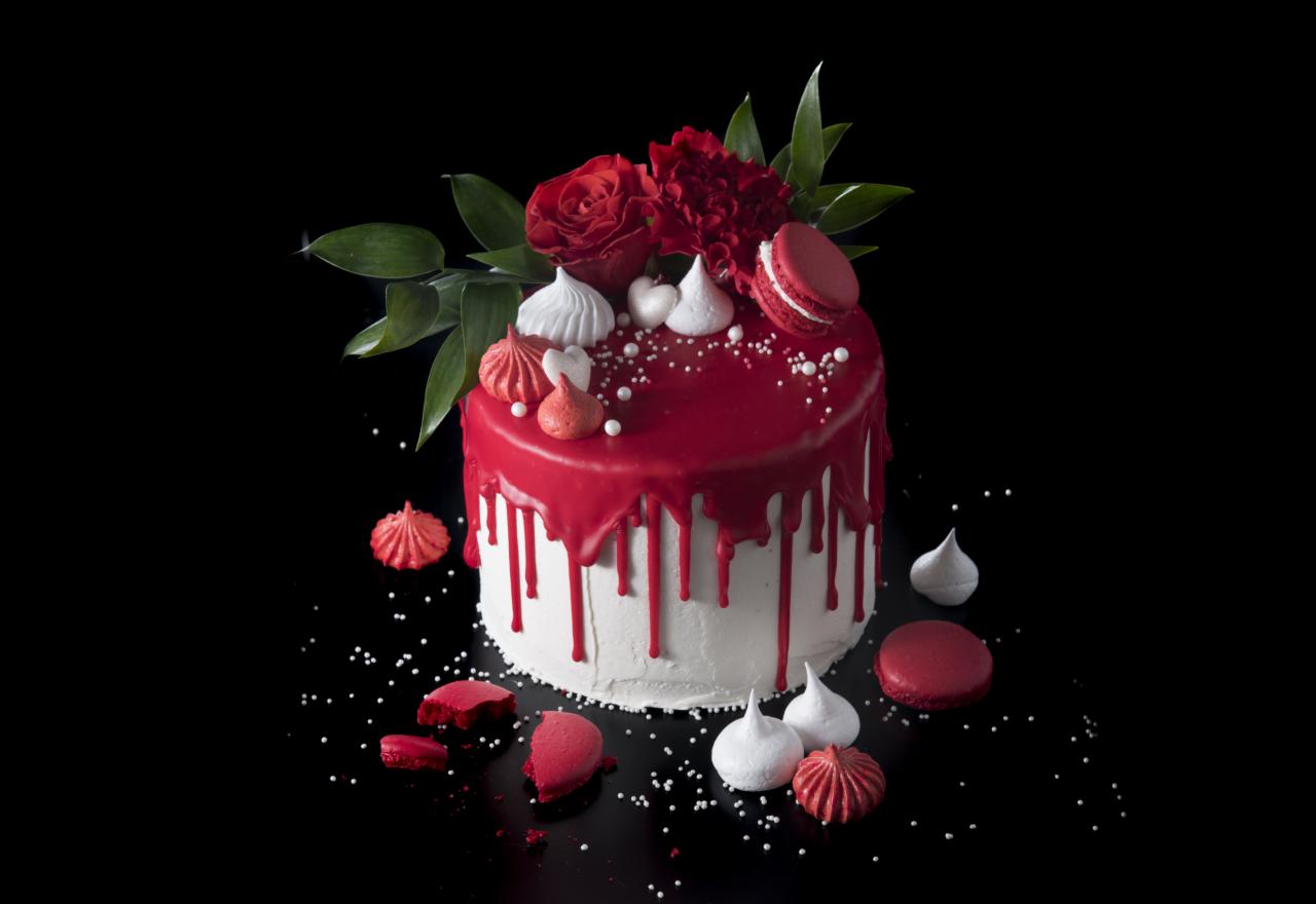 Tort z białym kremem, czerwonym lukrem, dekorowany czerwonymi makaronikami, bezami i żywymi czerwonymi kwiatami na czarnym tle.