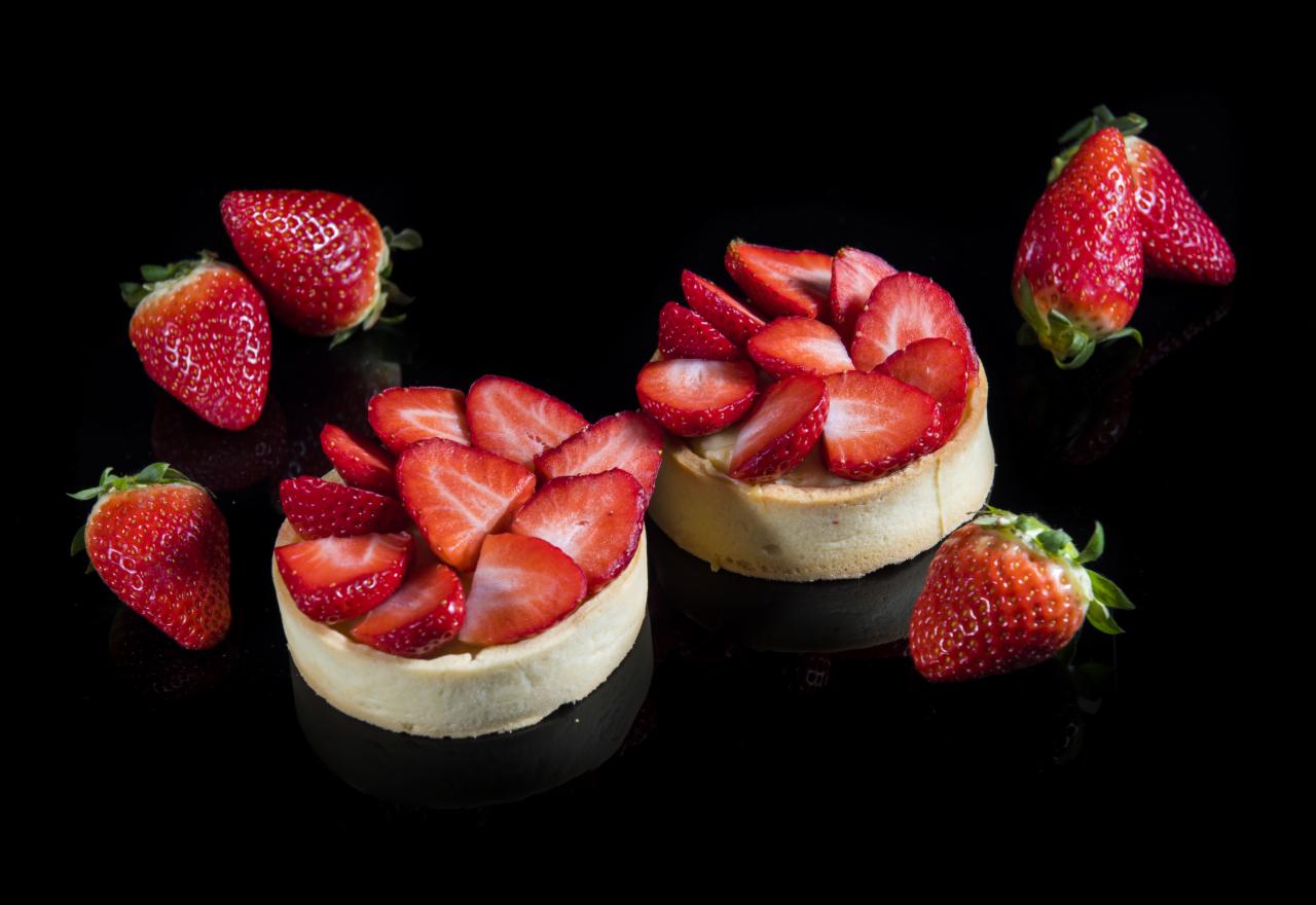 Dwie mini tartaletki z kremem i świeżymi truskawkami na błyszczącym czarnym tle, otoczone pojedynczymi truskawkami.