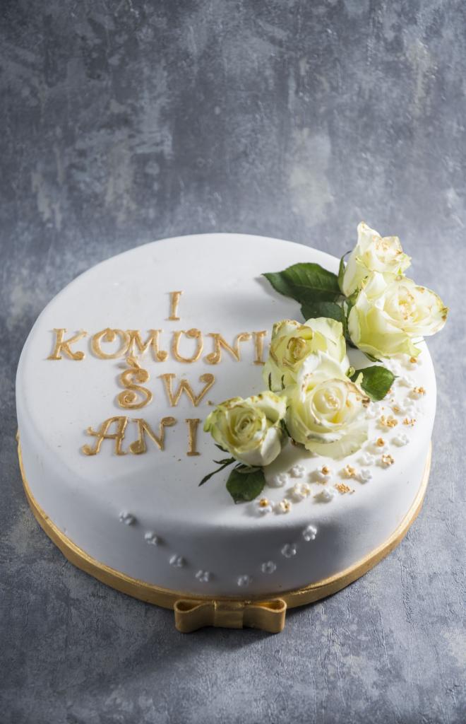 Tort okolicznościowy z białym lukrem, złotymi napisami 'I Komunii Św. Ani' i dekoracją z kremowych róż oraz złotych kuleczek, na szarym tle.
