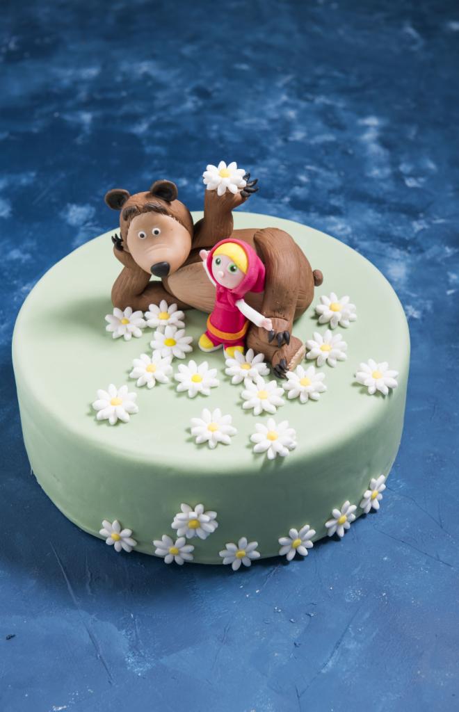 Tort zdobiony figurkami Masha i Niedźwiedź, z zielonym lukrem i białymi stokrotkami, na niebieskim tle.