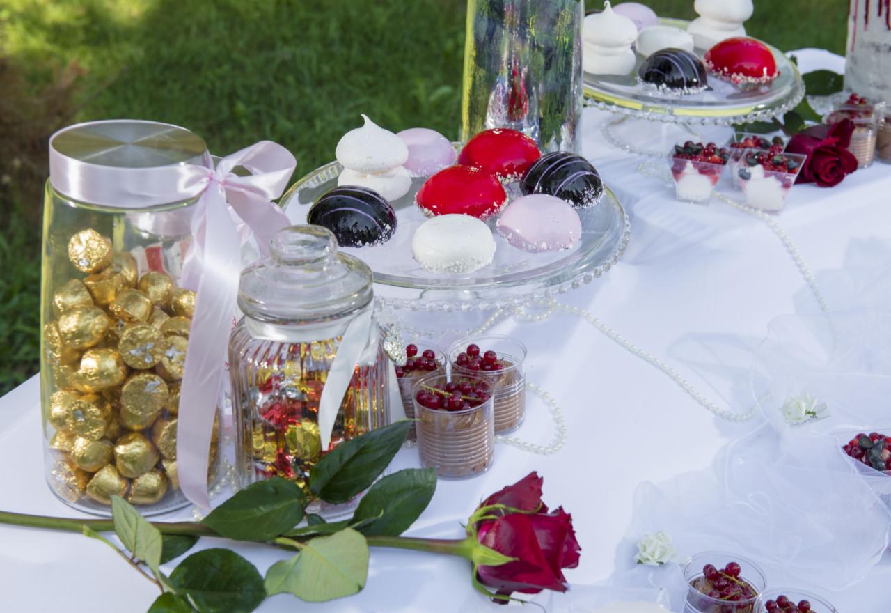 Słodki stół z deserami: błyszczące monoporcje, bezy, czekoladki w złotych papierkach w słoiku z różową wstążką, mini desery z owocami i róże na białym obrusie.