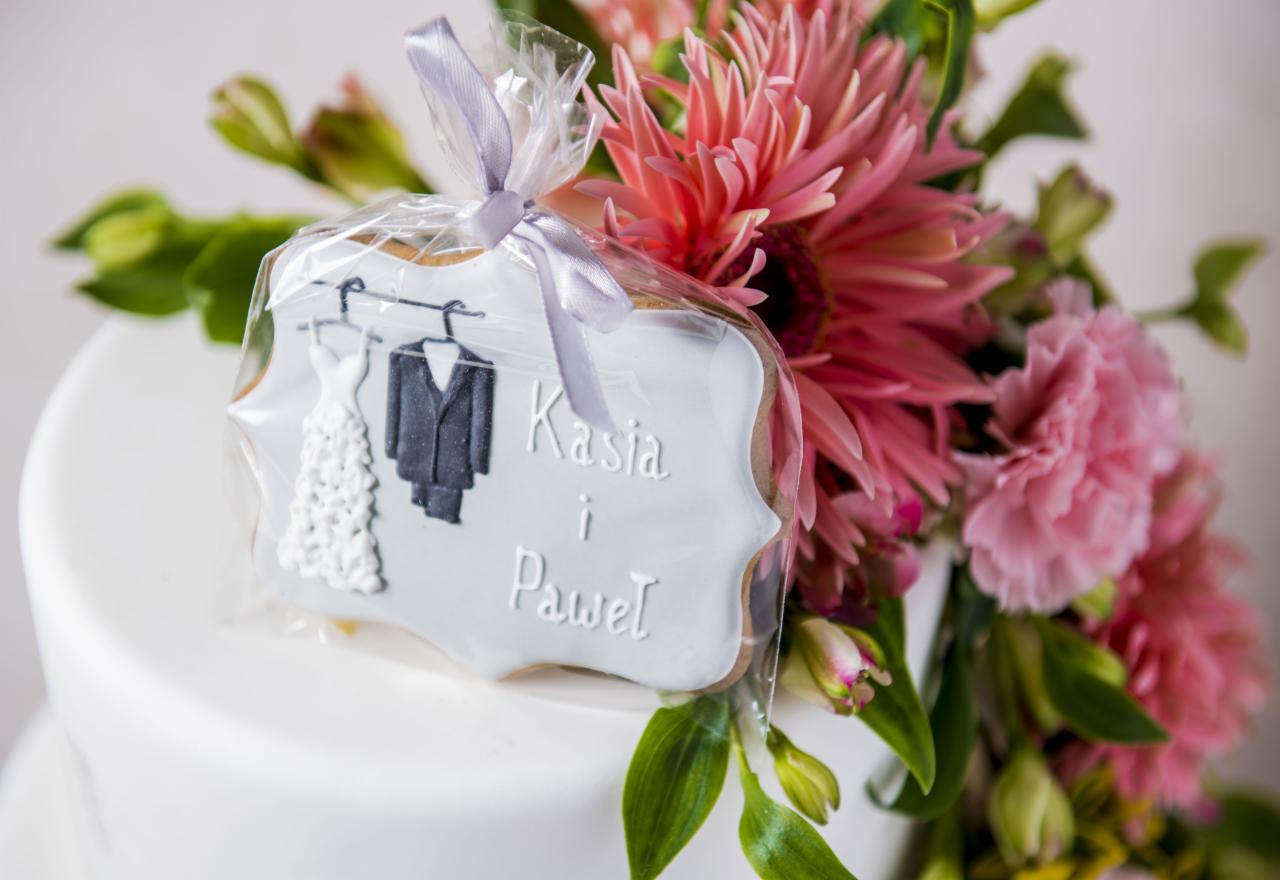 Piernik w kształcie ramki z imionami Kasia i Paweł, dekorowany motywem sukni ślubnej i garnituru, zapakowany w celofan i zawiązany wstążką, umieszczony na białym torcie z kwiatową dekoracją...