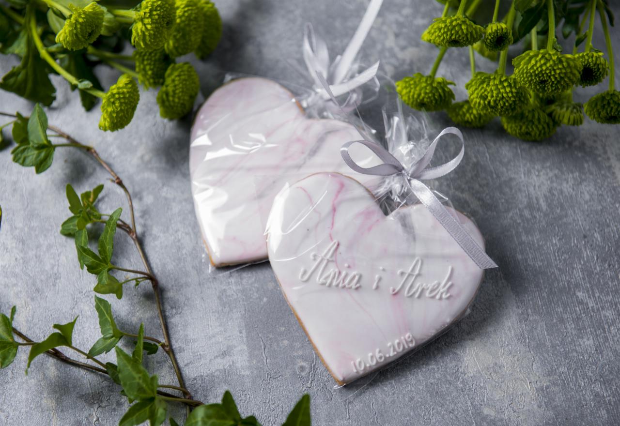 Dwa piernikowe serca w przezroczystych opakowaniach z kokardkami, z imionami 'Ania i Arek' oraz datą 10.06.2019, leżące na szarym tle z zielonymi kwiatami i gałązką bluszczu.