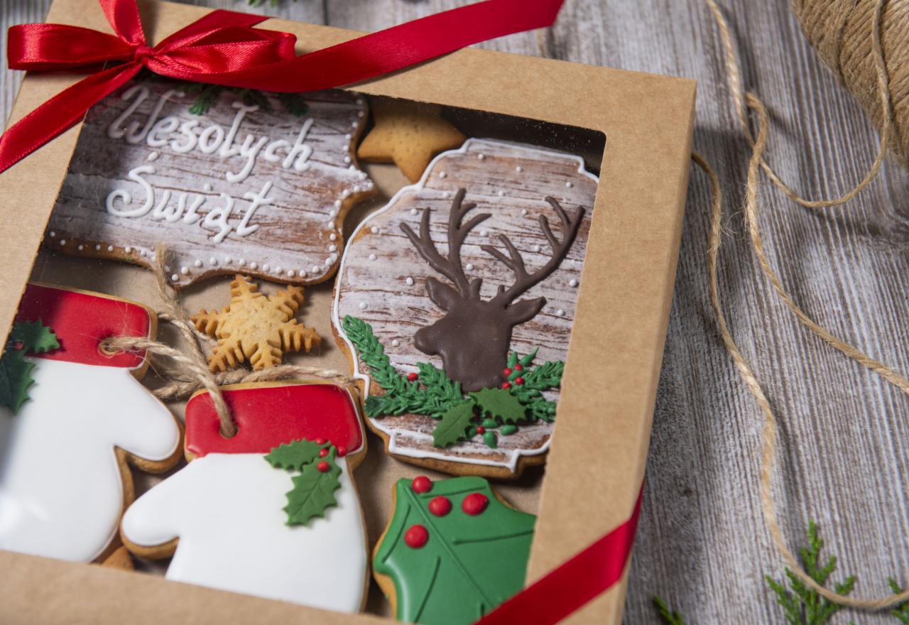 Pudełko z piernikami świątecznymi: renifer, rękawice, gwiazda i napis 'Wesołych Świąt', ozdobione lukrem i czerwoną wstążką, leżące na drewnianym tle.
