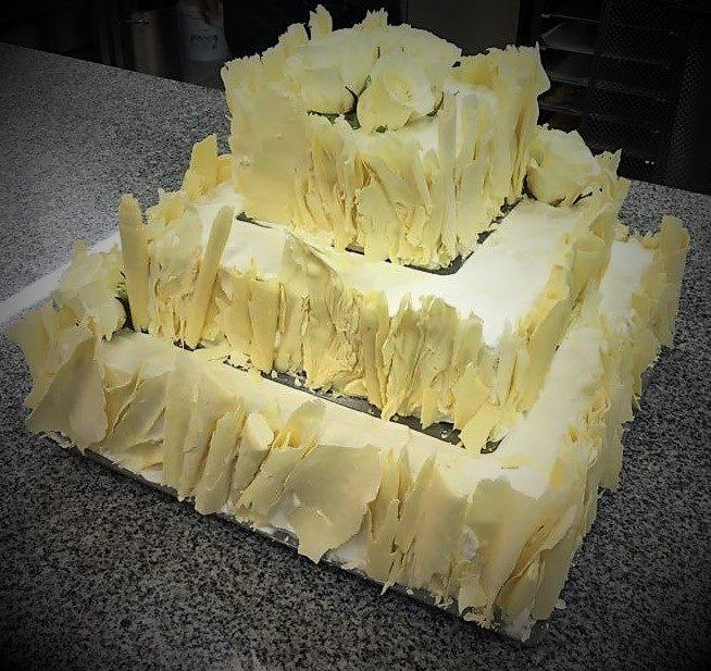 Trzypiętrowy tort kwadratowy, obłożony płatkami białej czekolady i ozdobiony białymi różami na szczycie, ustawiony na blacie z szarego granitu.