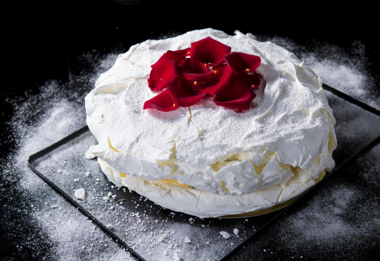 Tort bezowy Pavlova z płatkami róż na czarnym tle posypanym cukrem pudrem.