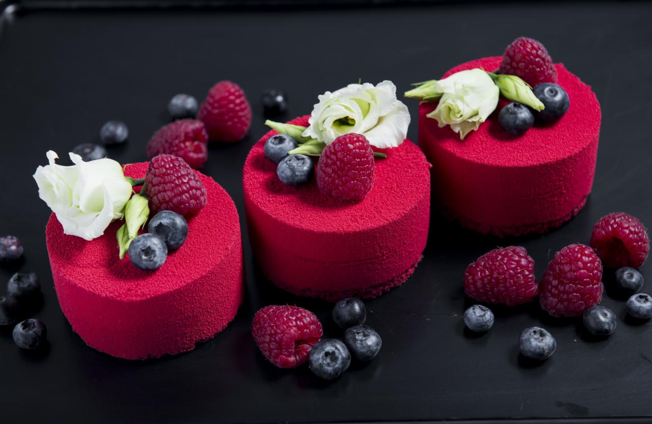 Trzy małe, okrągłe ciastka o intensywnie czerwonej, aksamitnej teksturze, udekorowane świeżymi malinami, borówkami i białymi kwiatami na czarnym tle.