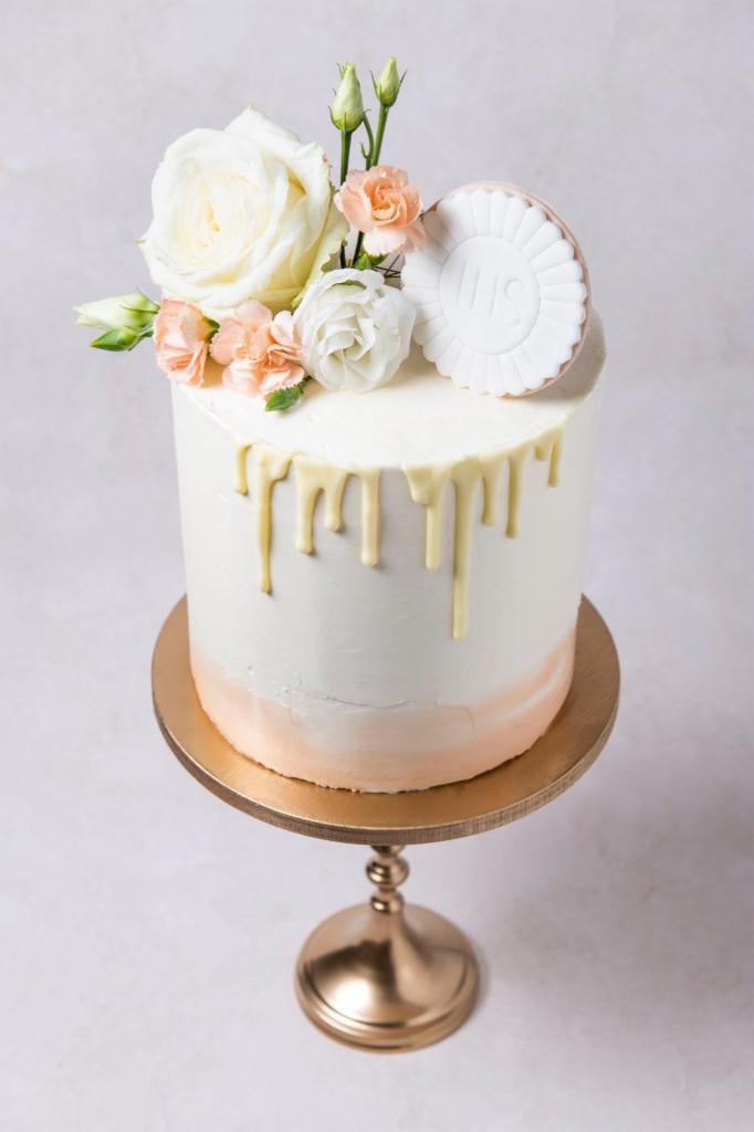 Elegancki tort komunijny z białym lukrem spływającym po bokach, ozdobiony białymi i brzoskwiniowymi różami oraz okrągłym ciasteczkiem z napisem IHS, umieszczony na złotej paterze.