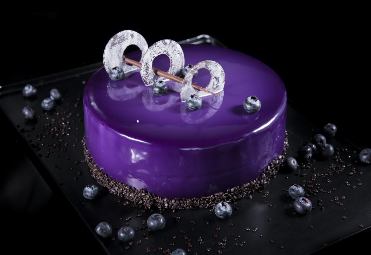 Lustrzany tort w odcieniach fioletu, udekorowany świeżymi borówkami i abstrakcyjnymi, białymi dekoracjami na czarnym tle, posypany drobnymi kawałkami czekolady.