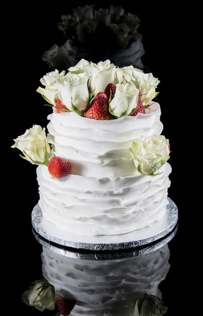 Dwupoziomowy tort weselny z białym kremem w falistej fakturze, udekorowany białymi różami i świeżymi truskawkami na czarnym, lustrzanym tle.