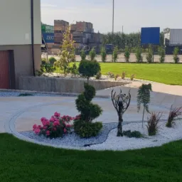 Elegancki ogród z trawnikiem, ścieżką z kostki, rabatami z białego kamienia i ozdobnymi roślinami. W tle budynek z garażem i zielenią.