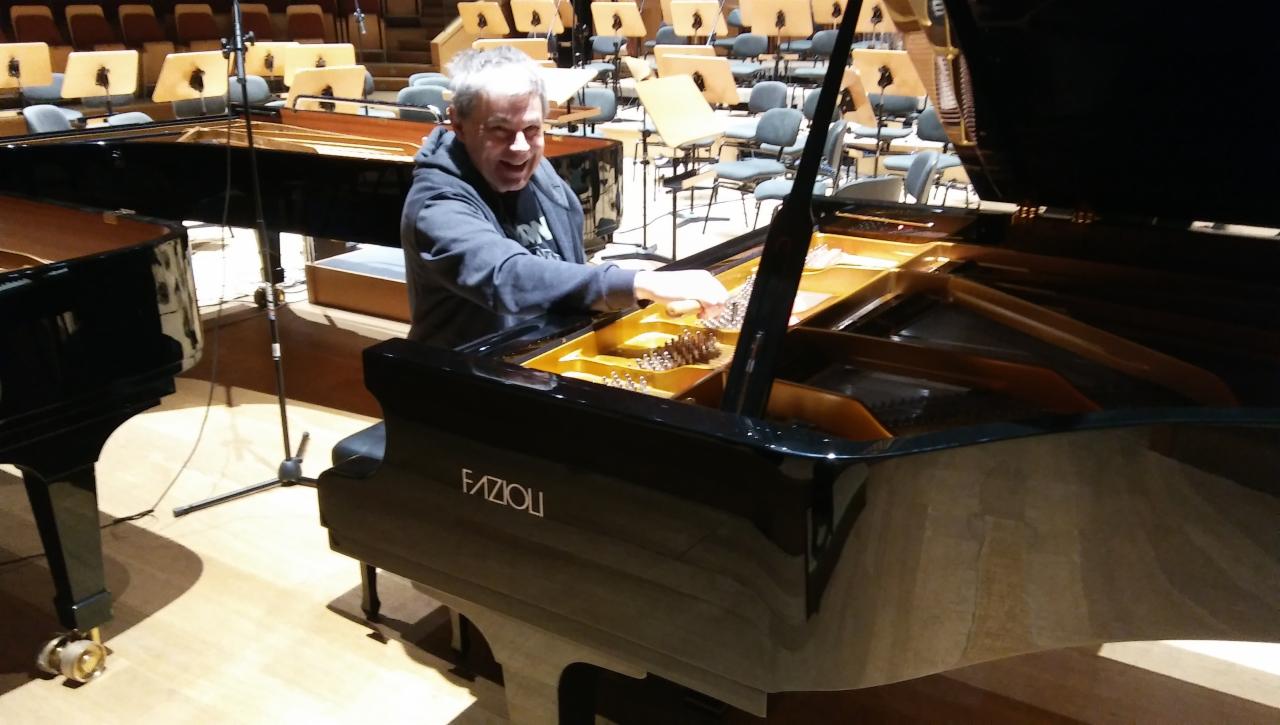 Mężczyzna w szarej bluzie z kapturem uśmiecha się grając na czarnym fortepianie Fazioli w sali koncertowej z rzędami pustych krzeseł w tle.