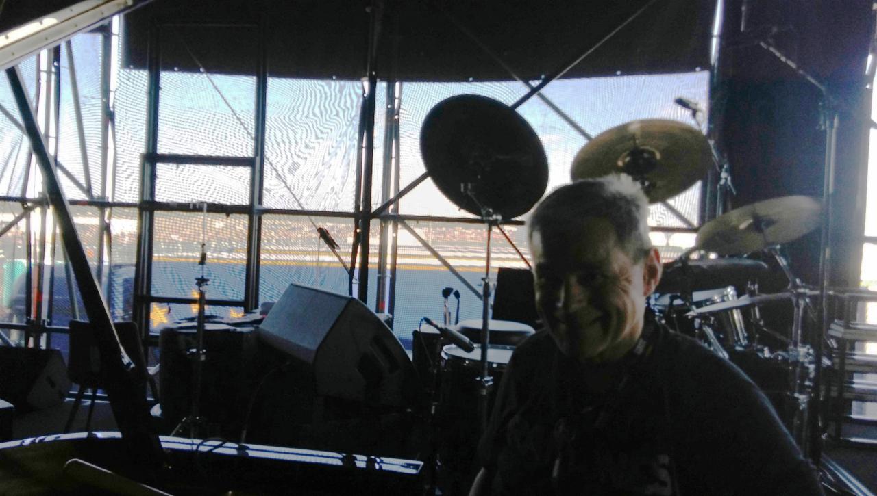 Uśmiechnięty perkusista na scenie przed koncertem, widoczny zestaw perkusyjny i nagłośnienie, tło stanowi duża przeszklona konstrukcja.