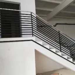 Białe schody z nowoczesną, czarno-srebrną balustradą z poziomych prętów, widoczne gniazdko elektryczne na ścianie i surowy betonowy strop.