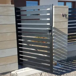 Nowoczesna, metalowa brama posesji z poziomymi szczeblami, zamontowana w Końskich, widoczna z zewnątrz, z fragmentem ogrodzenia z bloczk&oacute;w betonowych w tle.