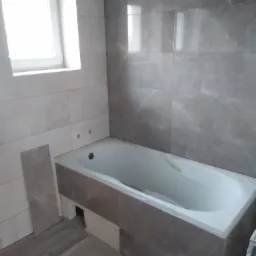 Łazienka w trakcie remontu: wanna obudowana płytkami imitującymi marmur, częściowo ułożone płytki ścienne i podłogowe, widoczne przygotowania do dalszych prac.