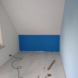 Pokój na poddaszu po malowaniu ścian na biało i niebiesko, z fragmentem pomalowanym na intensywny niebieski kolor, na podłodze leżą narzędzia: poziomica, miarka, ołówek i kabel od urządzenia.