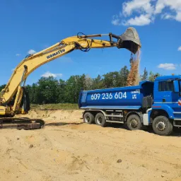 Żółta koparka Komatsu z uniesionym ramieniem wysypuje piasek do niebieskiej ciężarówki MAN z numerem telefonu na tle błękitnego nieba i zieleni drzew.
