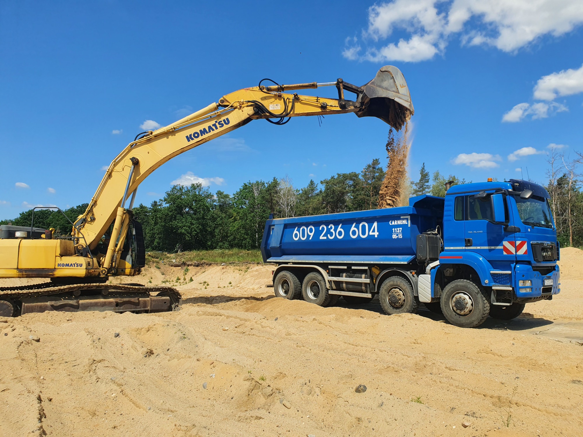 Żółta koparka Komatsu z uniesionym ramieniem wysypuje piasek do niebieskiej ciężarówki MAN z numerem telefonu na tle błękitnego nieba i zieleni drzew.