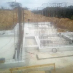 Wykonywanie fundamentów zbrojonych na rozległym placu budowy, widoczny pracownik w niebieskiej kurtce przy pracy, pionowe pręty zbrojeniowe wystające ponad poziom fundamentów.