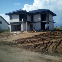 Dwukondygnacyjny dom w stanie surowym otwartym z białego bloczka, częściowo zadaszony, na placu budowy z błotnistym terenem, pod błękitnym niebem z białymi chmurami.