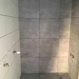 Wykończona łazienka z szarymi płytkami imitującymi beton na ścianach i podłodze, widoczne przyłącza wodne, minimalistyczny odpływ liniowy.