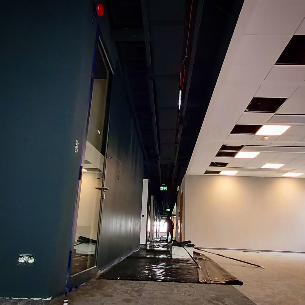 Wnętrze komercyjne w trakcie prac wykończeniowych: świeżo pomalowana ściana w odcieniach granatu, zabezpieczona folią podłoga, sufit kasetonowy z brakującymi elementami i widoczne instalacje...