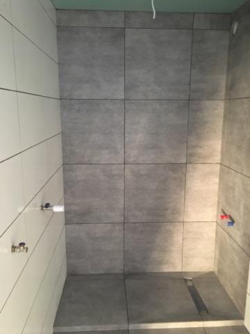 Wykończona łazienka z szarymi płytkami imitującymi beton na ścianach i podłodze, widoczne przyłącza wodne, minimalistyczny odpływ liniowy.