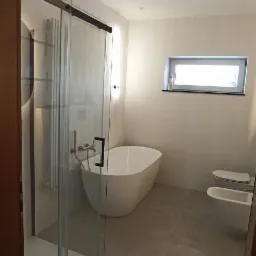 Nowoczesna łazienka z kabiną prysznicową typu walk-in z czarnymi detalami, wolnostojącą białą wanną, podwieszaną toaletą i bidetem na szarych płytkach podłogowych.