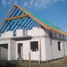 Surowy stan budowy domu jednorodzinnego z widoczną konstrukcją więźby dachowej, zabezpieczoną niebieską membraną, wykonaną na ścianach z betonu komórkowego, na tle błękitnego nieba.