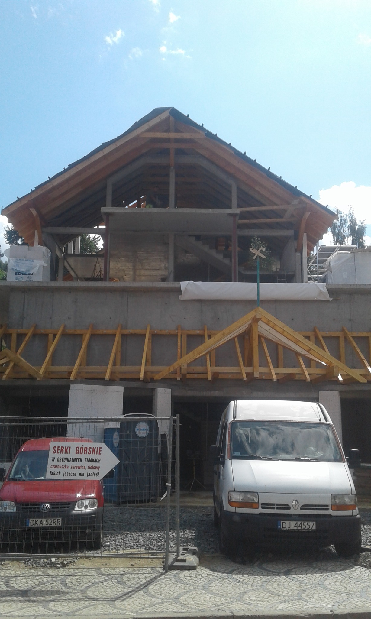 Surowa konstrukcja drewniana dachu i betonowego budynku w trakcie budowy, widoczne elementy więźby dachowej, szalunki, oraz pojazdy i tymczasowe ogrodzenie na pierwszym planie.