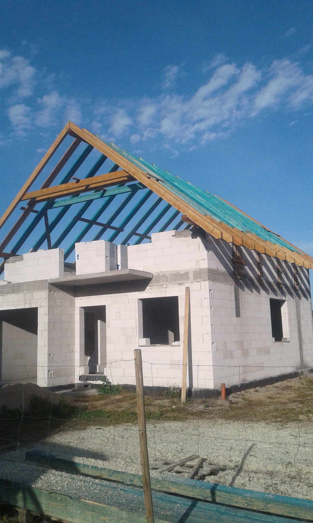 Surowy stan budowy domu jednorodzinnego z widoczną konstrukcją więźby dachowej, zabezpieczoną niebieską membraną, wykonaną na ścianach z betonu komórkowego, na tle błękitnego nieba.