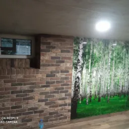 Ściana wykończona cegłą klinkierową z małym oknem z ozdobną kratą, sąsiadująca z fototapetą przedstawiającą brzozowy las, na podłodze panele imitujące drewno, widoczna butelka wody mineralnej.