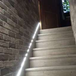 Schody z jasnego drewna z podświetleniem LED wzdłuż ceglanej ściany, prowadzące do otwartych drzwi.