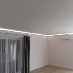 Wnętrze pokoju z oświetleniem LED w listwach przysufitowych, widoczne zwisające kable, klimatyzator na ścianie, szare zasłony przy oknie i panele podłogowe.