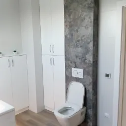 Nowoczesna toaleta podwieszana z szarą płytką dekoracyjną z motywem kwiatowym, białe szafki łazienkowe i drewnopodobna podłoga, widoczne przewody elektryczne.