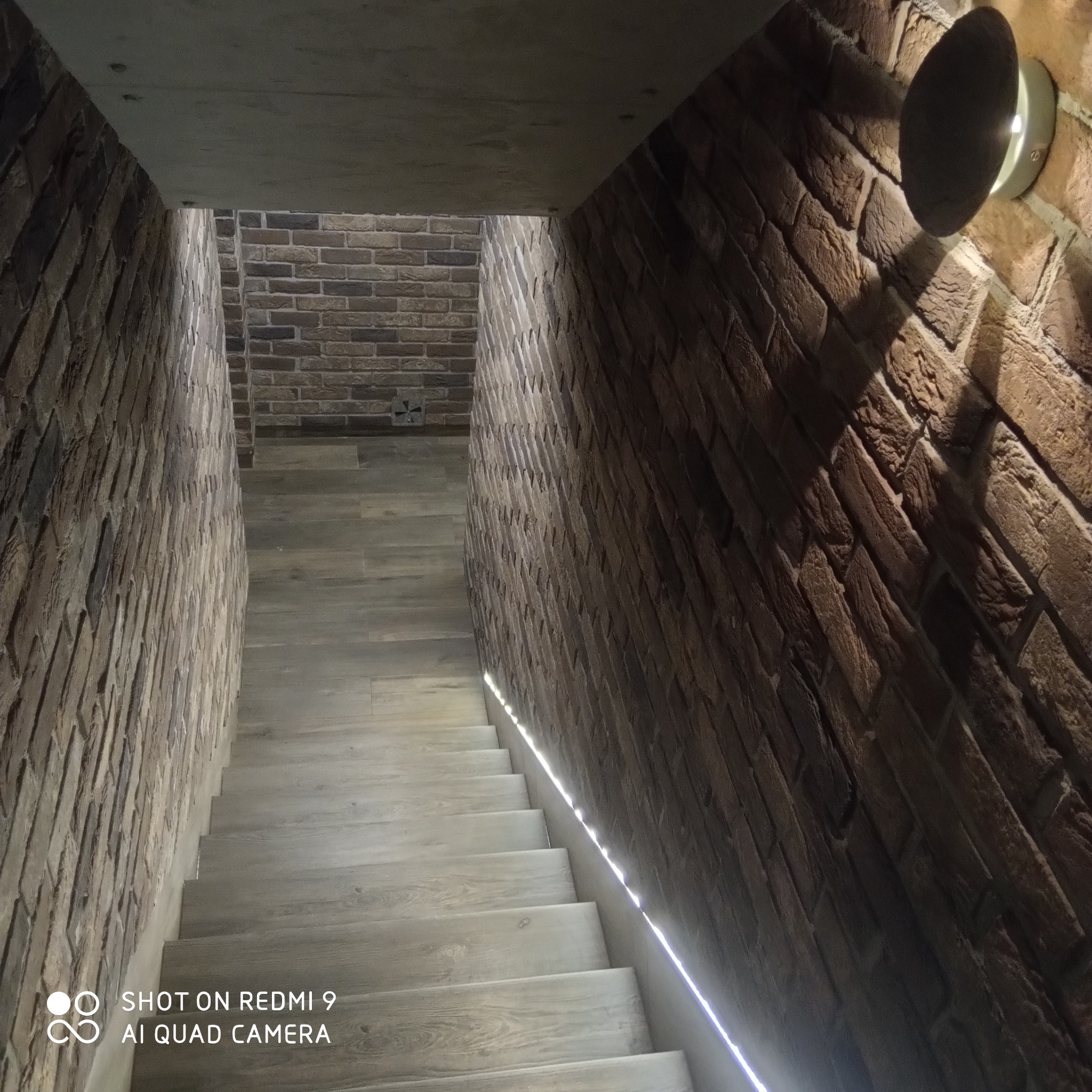 Klatka schodowa z drewnopodobnymi stopniami i ścianami wyłożonymi cegłą, oświetlona lampą ścienną i subtelnym paskiem LED. Widoczny fragment sufitu z betonu.