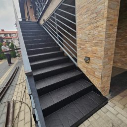 Uretanowe M Sp. z o. o. - Zewnętrzne schody z czarną, teksturowaną nawierzchnią żywiczną, z metalową balustradą. Widoczna elewacja budynku z kamienia dekoracyjnego i fragment chodnika z kostki brukowej.