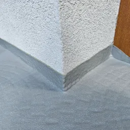 Szara, gładka posadzka żywiczna przy ścianie budynku z widocznym narożnikiem i fragmentem drzwi. Widoczne detale struktury posadzki i ściany.