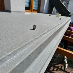 Szara, teksturowana powierzchnia z bliska, prawdopodobnie ocieplenie pianką PUR na dachu, z widocznymi elementami montażowymi i fragmentem rusztowania w tle.