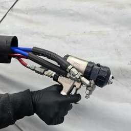 Uretanowe M Sp. z o. o. - Pistolet natryskowy z przewodami (czerwony, niebieski) w dłoni w czarnej rękawicy, gotowy do aplikacji na białą powierzchnię. Detal urządzenia i węży.