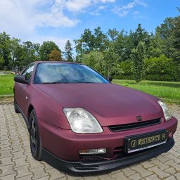 Uretanowe M Sp. z o. o. - Bordowy samochód Honda Prelude z unikalną, teksturowaną powłoką, zaparkowany na kostce brukowej w otoczeniu zieleni i błękitnego nieba. Widoczna tablica z napisem 'URETANOWE M'.