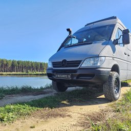 Uretanowe M Sp. z o. o. - Szary van Mercedes Sprinter z chropowatą powłoką ochronną, snorkel i opony terenowe, zaparkowany na szutrowej drodze z widokiem na jezioro i las.