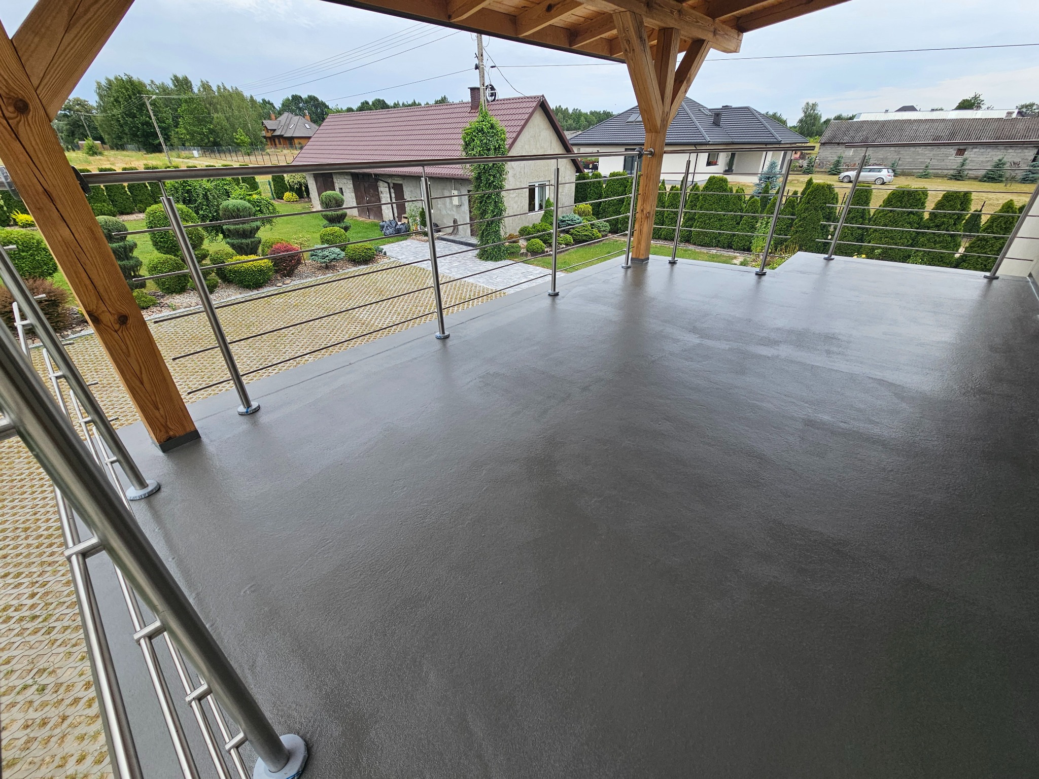 Szara posadzka żywiczna na balkonie z drewnianą konstrukcją i metalową balustradą. W tle widok na zadbany ogród i domy.