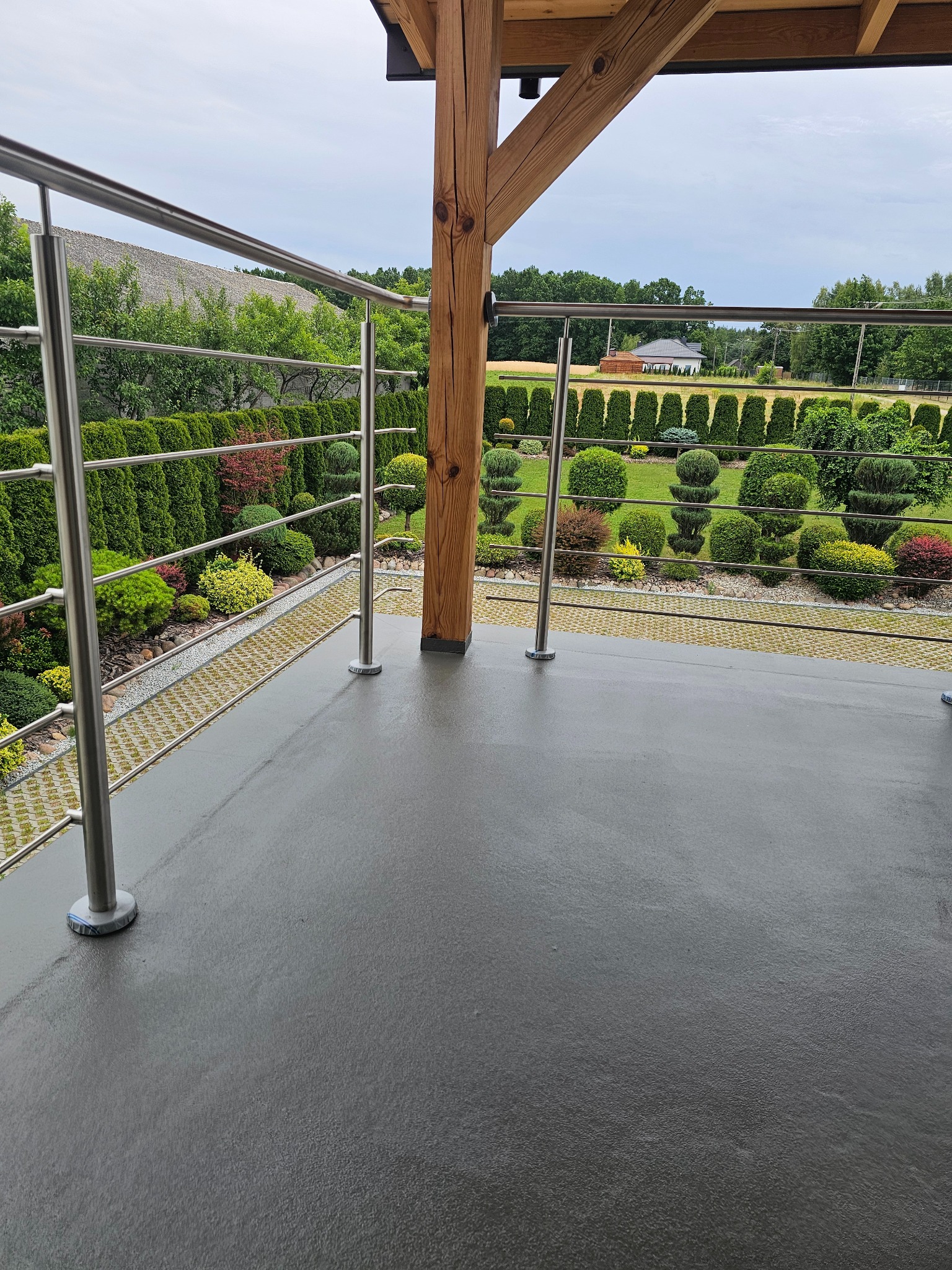 Szara, gładka posadzka żywiczna na balkonie z metalową balustradą i widokiem na zadbany ogród z formowanymi krzewami. Drewniany element konstrukcyjny w tle.