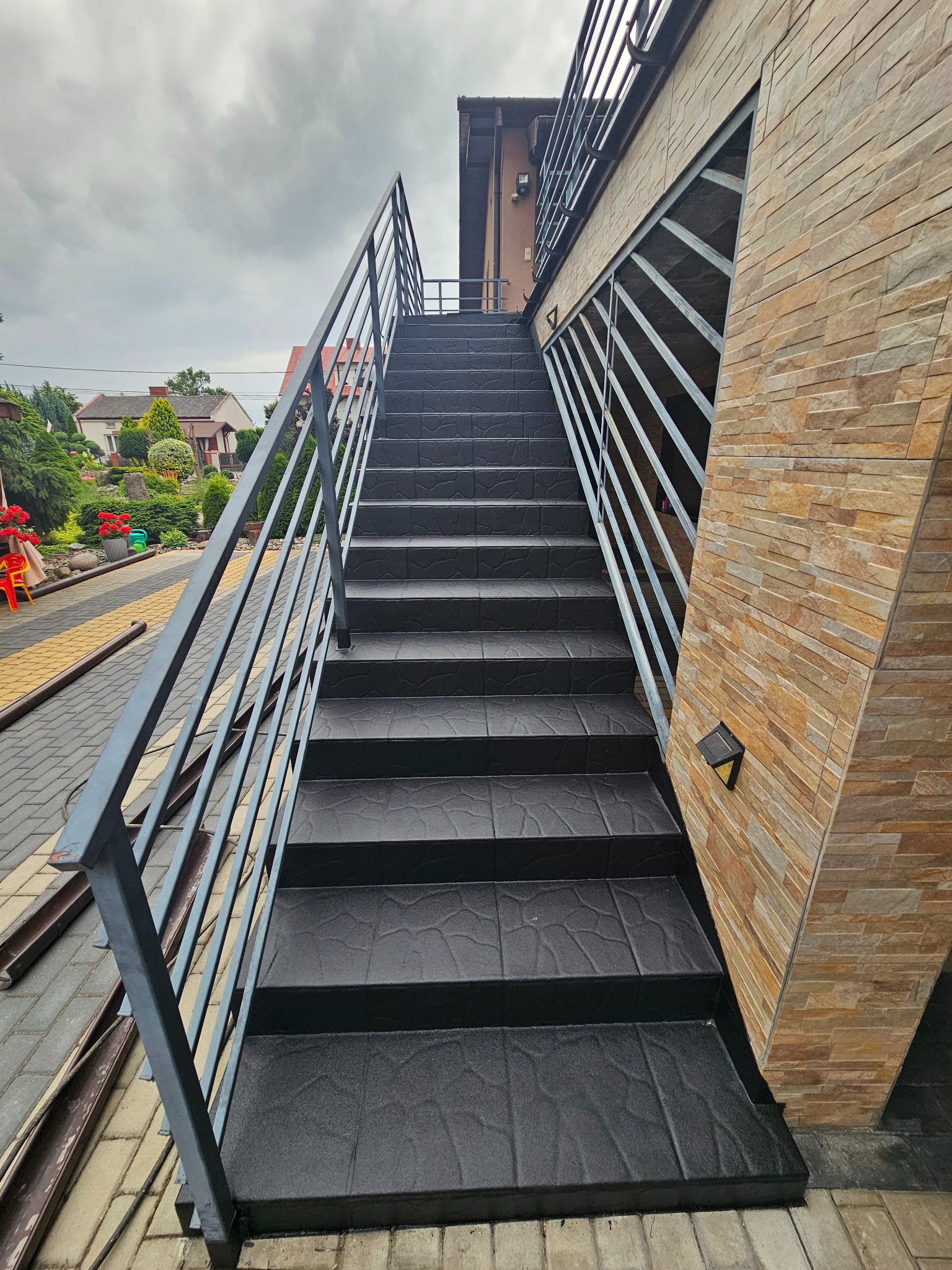 Zewnętrzne schody z czarną, wzorzystą posadzką żywiczną i metalową balustradą, przylegające do elewacji z kamienia dekoracyjnego w odcieniach beżu.