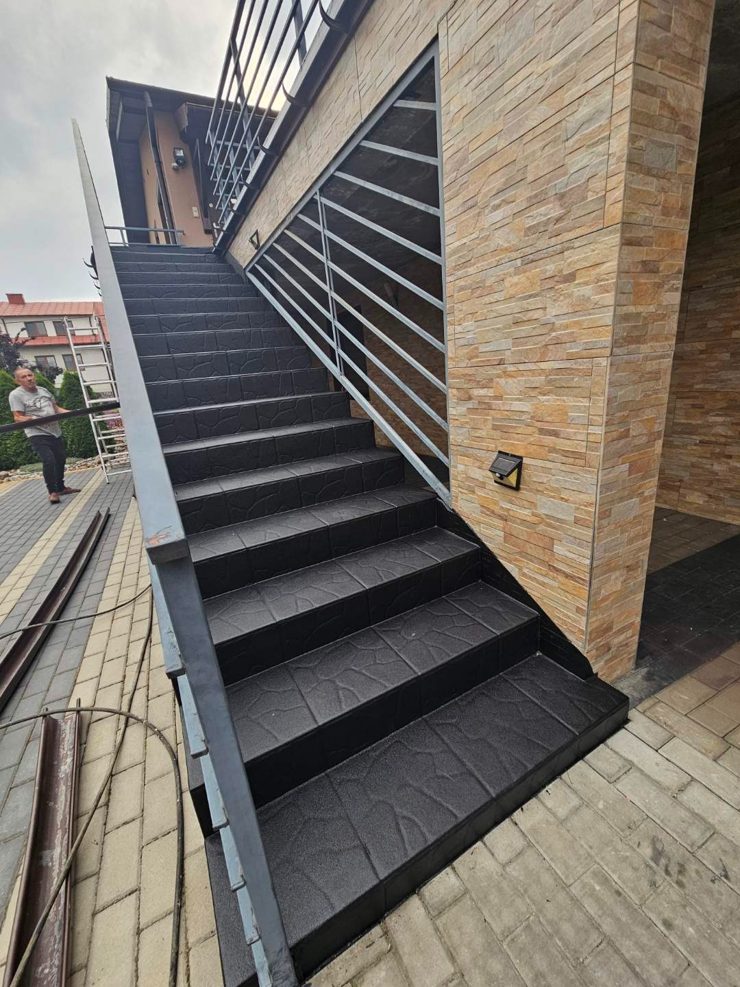 Zewnętrzne schody z czarną, teksturowaną nawierzchnią żywiczną, z metalową balustradą. Widoczna elewacja budynku z kamienia dekoracyjnego i fragment chodnika z kostki brukowej.