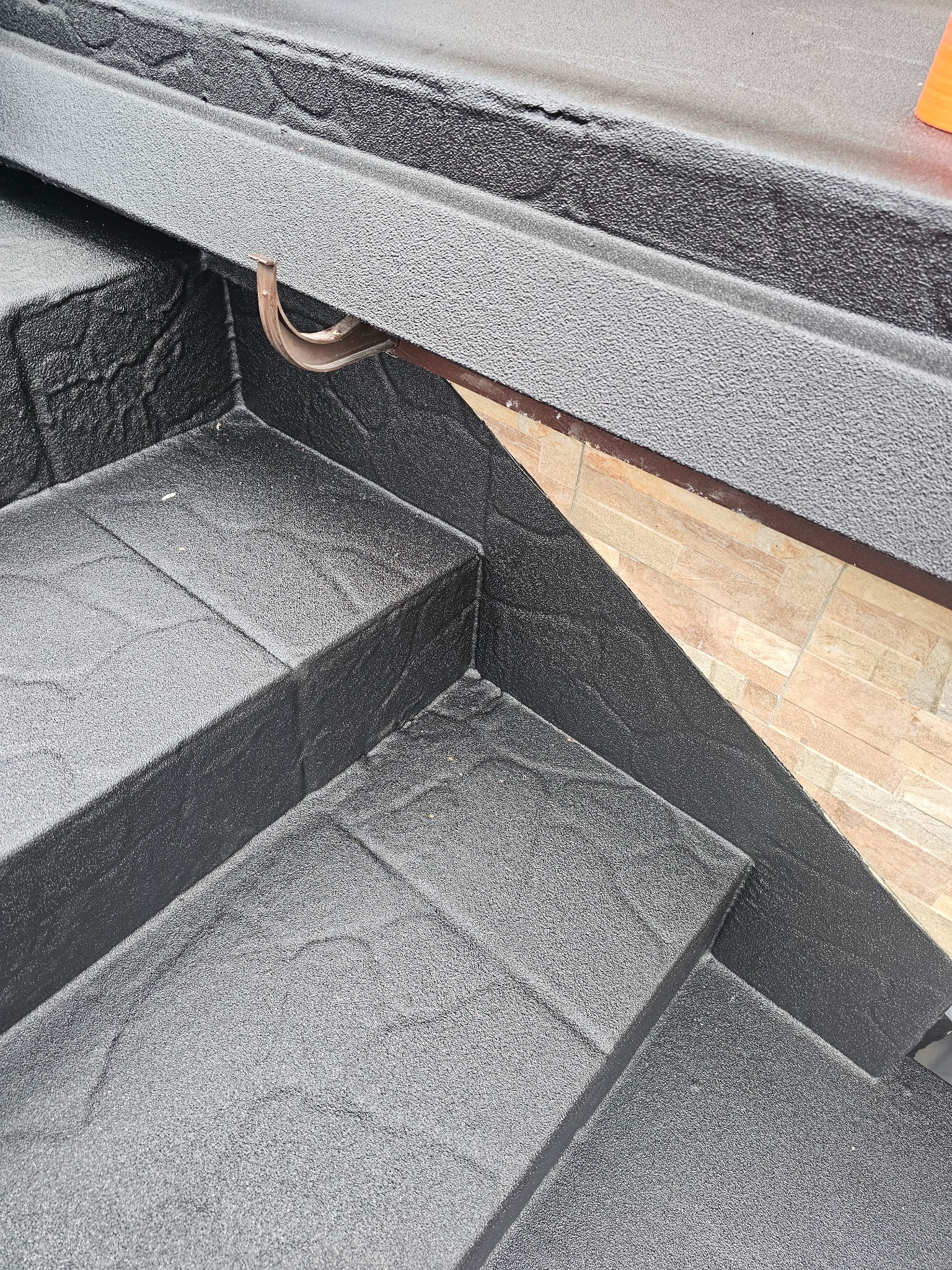 Schody zewnętrzne pokryte czarną, chropowatą posadzką żywiczną, z widocznym fragmentem brązowej rynny i okładziny ściennej z płytek imitujących cegłę.